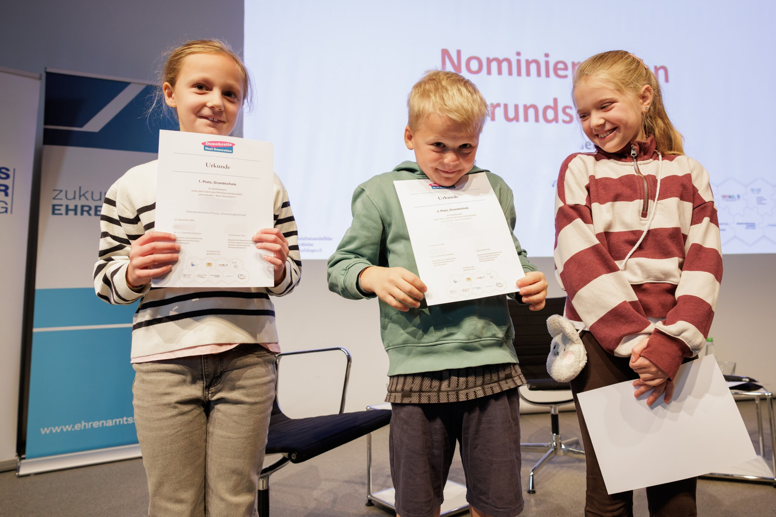 Ein Schüler und zwei Schülerinnen posieren mit ihren Urkunden auf der Bühne bei der Preisverleihung des Projekts „Demokratie – Next Generation“.