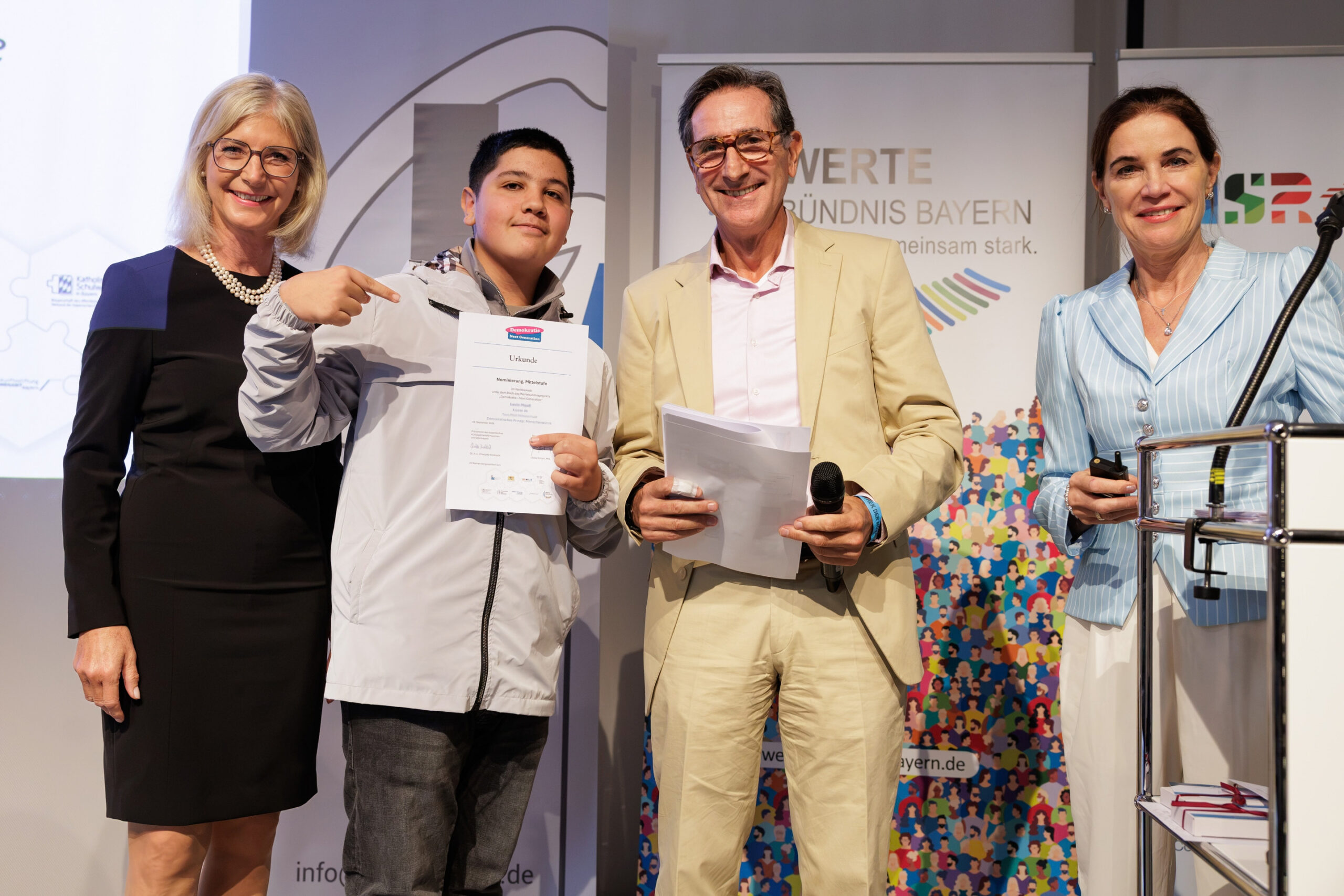 Ein Schüler posiert mit seiner Urkunde, gemeinsam mit Ulrike Scharf, MdL, Yehoshua Chmiel und Karin Schnebel auf der Bühne bei der Preisverleihung des Projekts „Demokratie – Next Generation“.