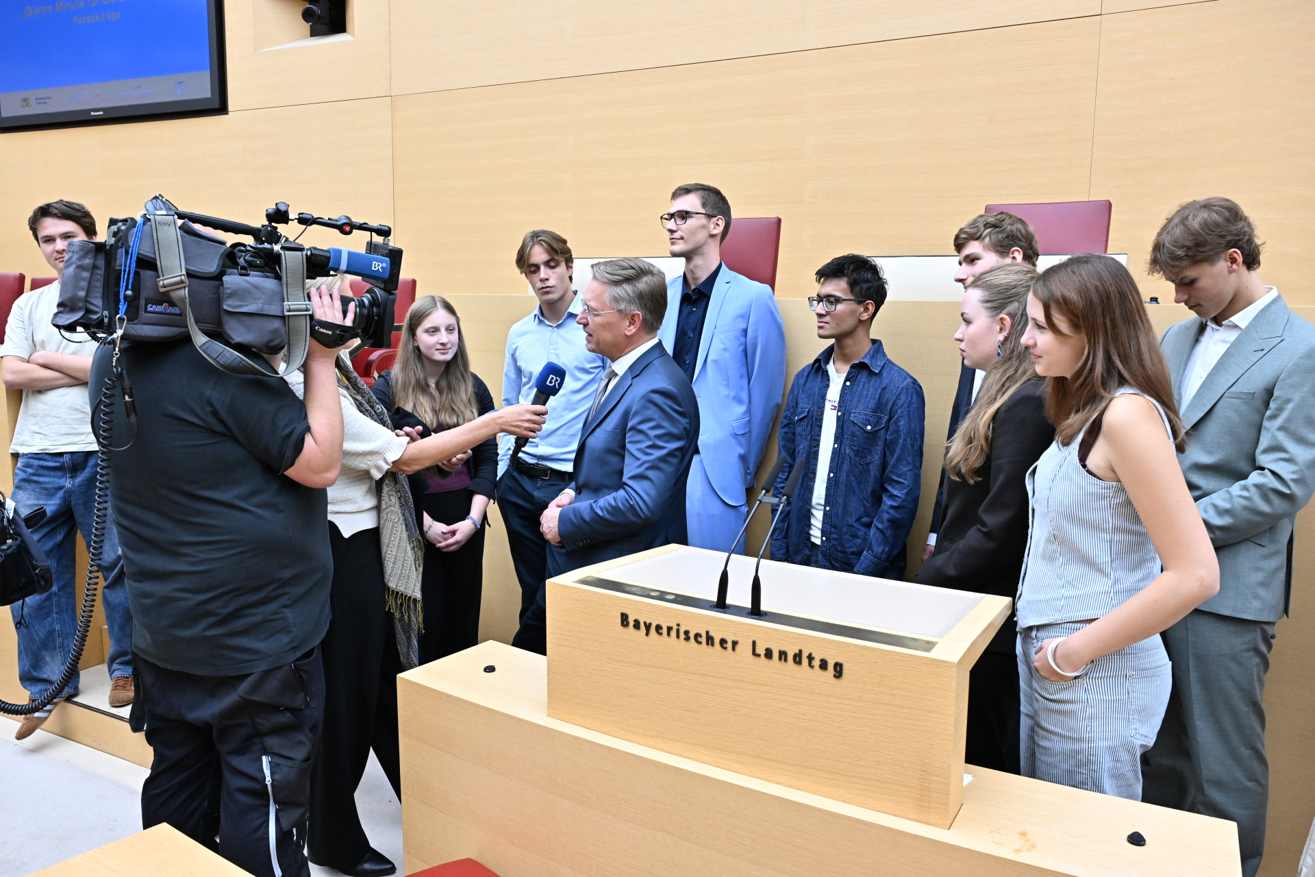 Vizepräsident des Bayerischen Landtags, Tobias Reiss gibt ein Interview nach dem Projekt „(D)eine Minute für die Demokratie", umringt von den jungen Rednerinnen und Rednern.